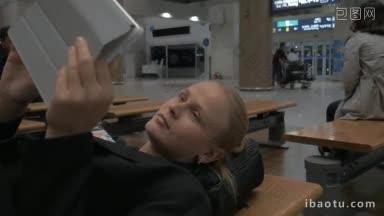疲倦的女士躺在机场休息室的<strong>长凳</strong>上，用平板电脑等飞机